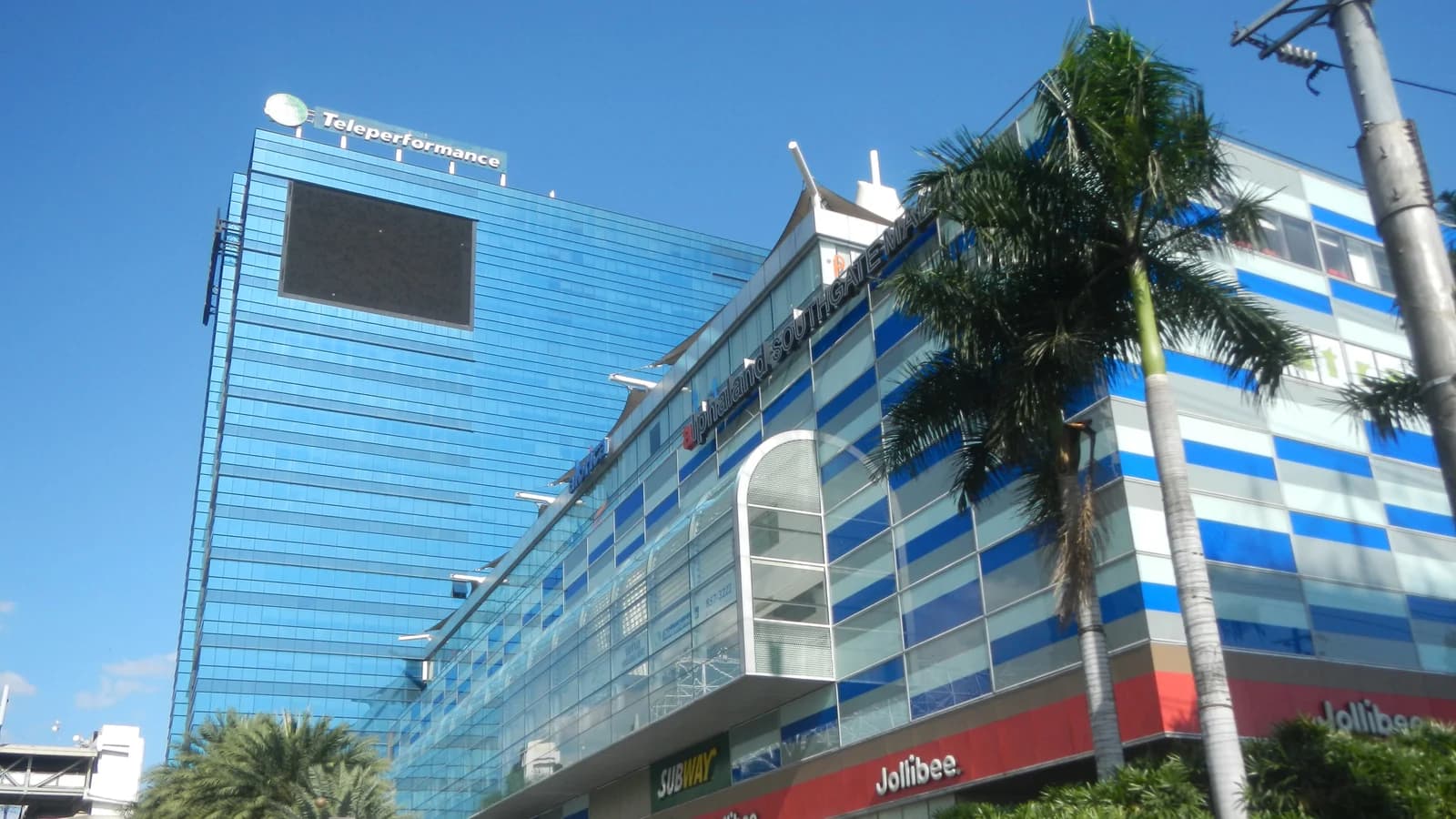 Gedung kantor Teleperformance di Makati, Filipina: menara kaca biru dengan logo Teleperformance di puncaknya, di samping mal dan pohon palem.